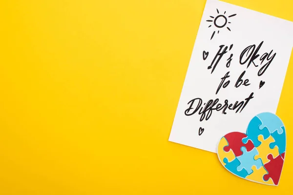 Top view of paper sheet with its okay to be different inscription  and puzzle heart on yellow — Stock Photo