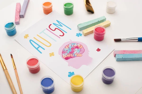 Card with autism word and painting of head with brain and puzzle on white with paint brushes, chalks and paints — Stock Photo