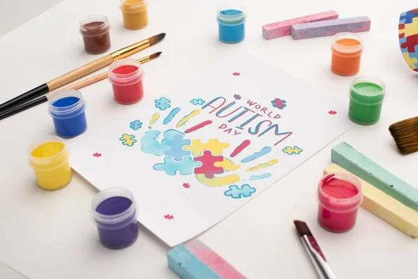 Card with World Autism Day lettering and painting of puzzle and hand prints on white with paint brushes, chalks and paints — Stock Photo