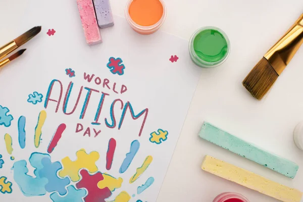 Vista superior de la tarjeta con letras del Día Mundial del Autismo y pintura de rompecabezas y huellas de mano en blanco con pinceles, tiza y pinturas - foto de stock