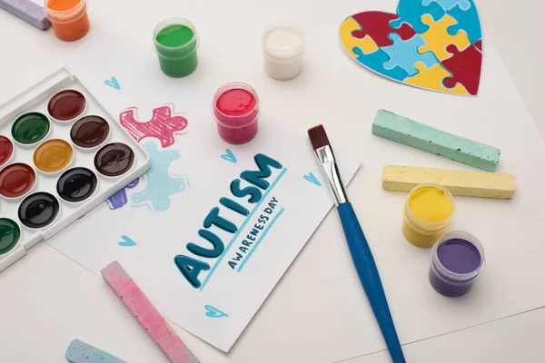 Tarjeta con Autism Awareness Day letras y pintura de rompecabezas en blanco con pinceles, tiza y corazón del rompecabezas - foto de stock