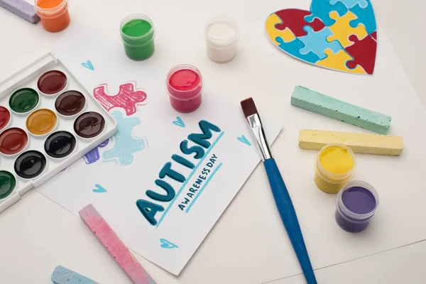 Tarjeta con Autism Awareness Day letras y pintura de rompecabezas en blanco con pinceles, tiza y corazón del rompecabezas — Stock Photo