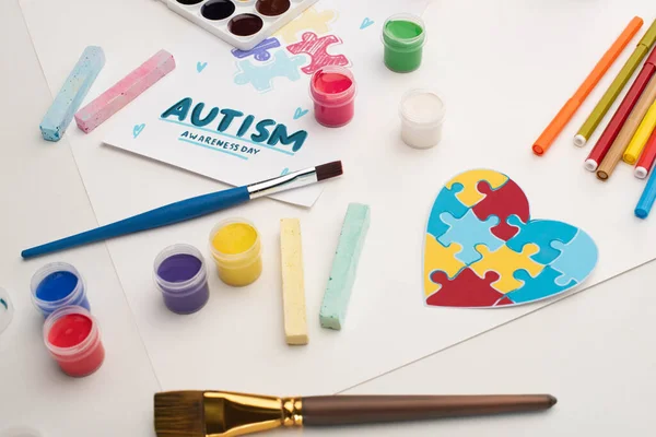 Card with Autism Awareness Day lettering and painting of puzzle on white with paint brushes, chalks, markers and puzzle heart — Stock Photo