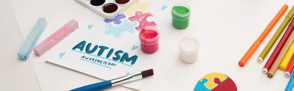 Panoramic shot of card with Autism Awareness Day lettering and painting of puzzle on white with paint brushes, chalks and markers — Stock Photo