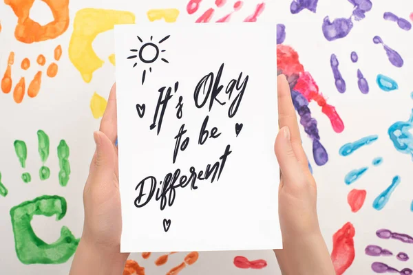 Cropped view of woman holding card with Its Okay to be different lettering on white with hand prints — Stock Photo