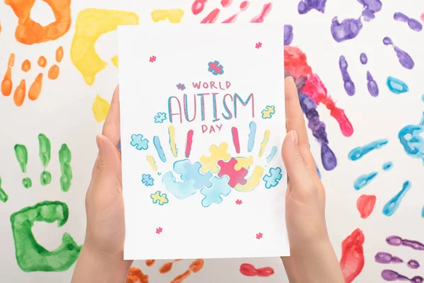 Cropped view of woman holding card with World Autism Day lettering and puzzle on white with hand prints — Stock Photo