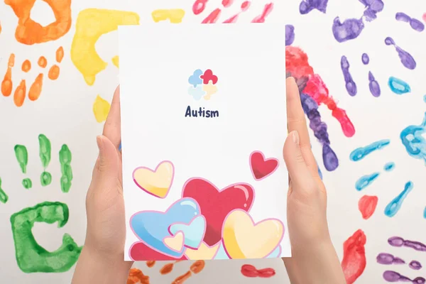 Vista recortada de la mujer que sostiene la tarjeta con la palabra autismo y corazones en blanco con impresiones de mano - foto de stock