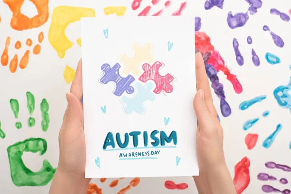 Cropped view of woman holding card with Autism Awareness Day lettering and puzzle on white with hand prints — Stock Photo