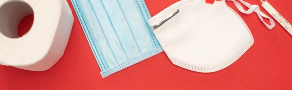 Top view of white toilet paper, safety and medical masks and thermometer on red background, panoramic shot — Stock Photo