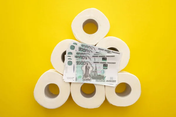 Vista dall'alto di rotoli di carta igienica bianca disposti in piramide con soldi russi su sfondo giallo — Foto stock