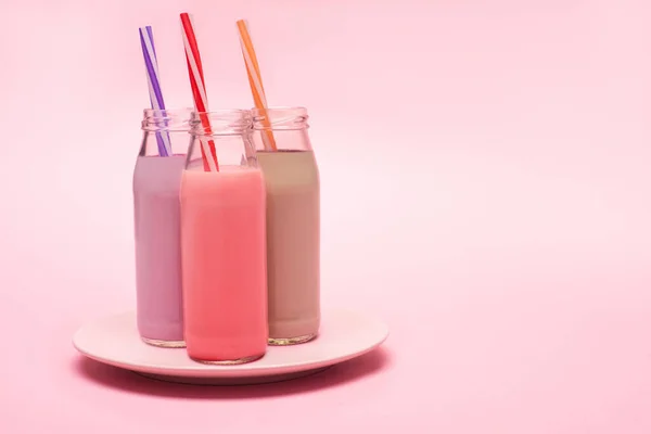 Bottles of berry, strawberry and chocolate milkshakes with drinking straws on plate on pink background — Stock Photo