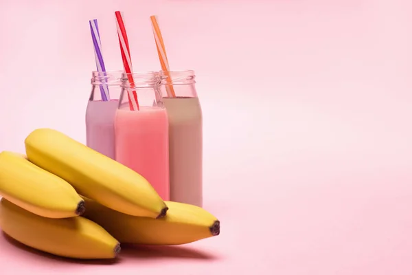 Botellas de batidos de bayas, fresas y chocolate con pajitas y plátanos sobre fondo rosa - foto de stock