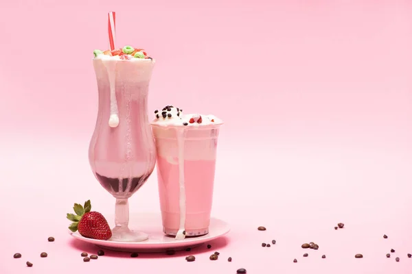 Tasse jetable et verre de milkshakes avec fraise sur assiette et grains de café sur fond rose — Photo de stock