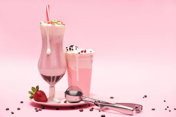 Copo descartável e vidro de milkshakes, morango e colher em prato com grãos de café em rosa — Fotografia de Stock