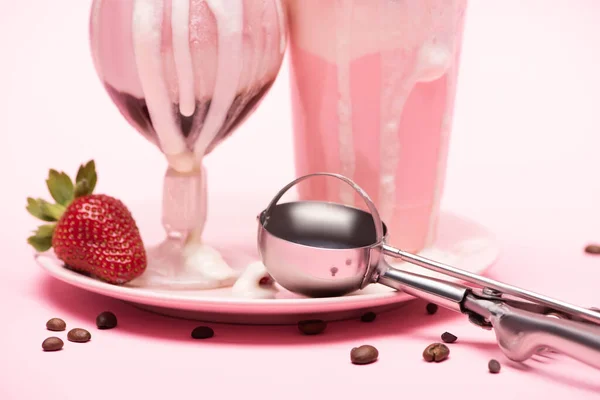 Tasse jetable et verre de milkshakes, fraise et cuillère sur plaque avec grains de café sur fond rose — Photo de stock