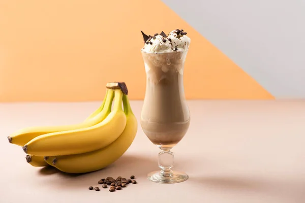 Vaso de batido con trozos de chocolate cerca de plátanos y granos de café en beige, gris y naranja - foto de stock