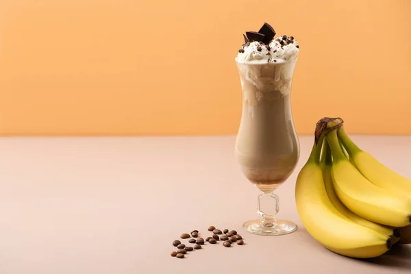 Vaso de batido con trozos de chocolate cerca de plátanos y granos de café en beige y naranja - foto de stock