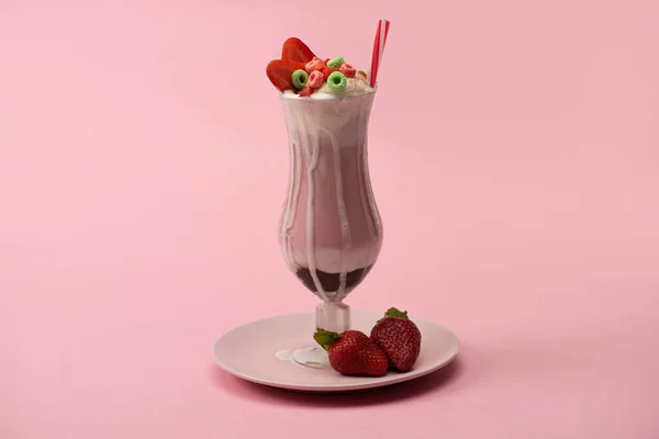Склянка молочного коктейлю з питною соломою, цукерками та полуницею на тарілці на рожевому фоні — стокове фото