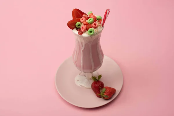 High angle view of glass of milkshake with drinking straw, candies and strawberries on plate on pink background — Stock Photo