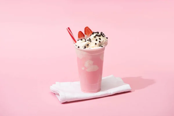 Disposable cup of milkshake with drinking straw, chocolate chips and strawberry halves on napkins on pink — Stock Photo