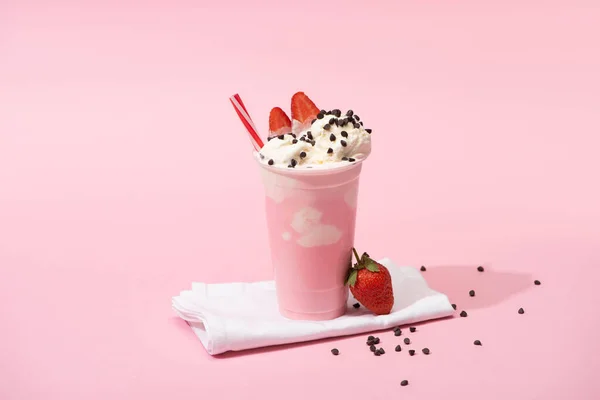 Disposable cup of milkshake with drinking straw, chocolate chips and strawberries on napkins on pink background — Stock Photo