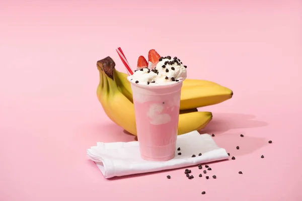 Disposable cup of strawberry milkshake with chocolate chips on napkins near bananas on pink background — Stock Photo