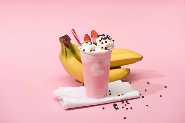 Einwegbecher Erdbeer-Milchshake mit Schokoladenchips auf Servietten in der Nähe von Bananen auf rosa Hintergrund — Stockfoto