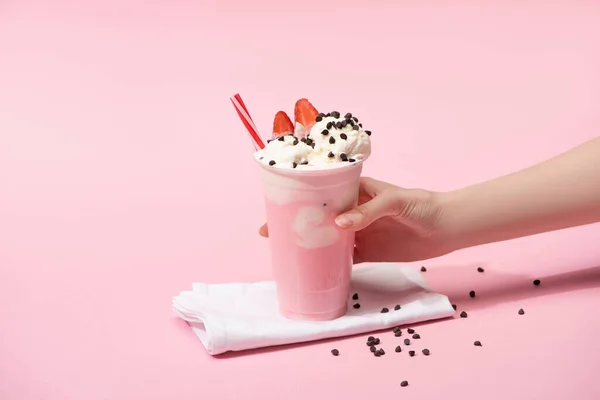 Обрезанный вид женской руки с одноразовой чашкой молочного коктейля с шоколадными кусочками и клубникой на салфетках на розовом — стоковое фото