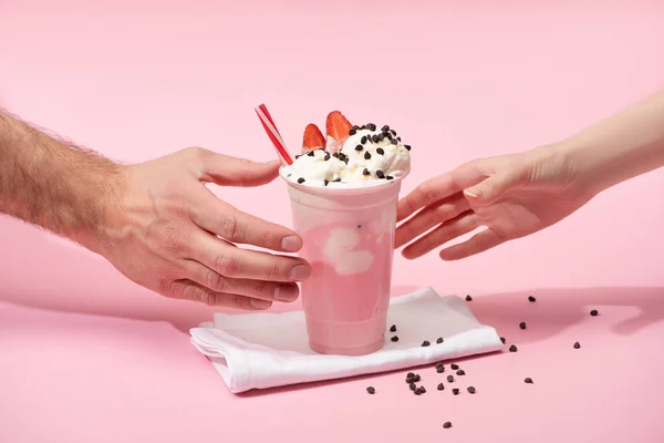 Partial view of female and male hands with disposable cup of strawberry milkshake with chocolate morsels on napkins on pink — Stock Photo