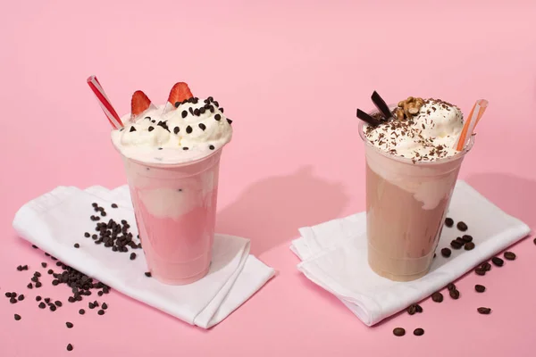 Disposable cups of chocolate and strawberry milkshakes with coffee grains on napkins on pink background — Stock Photo