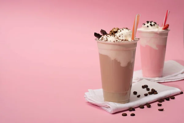 Enfoque selectivo de tazas desechables de batidos de chocolate y fresa con granos de café en servilletas sobre fondo rosa - foto de stock