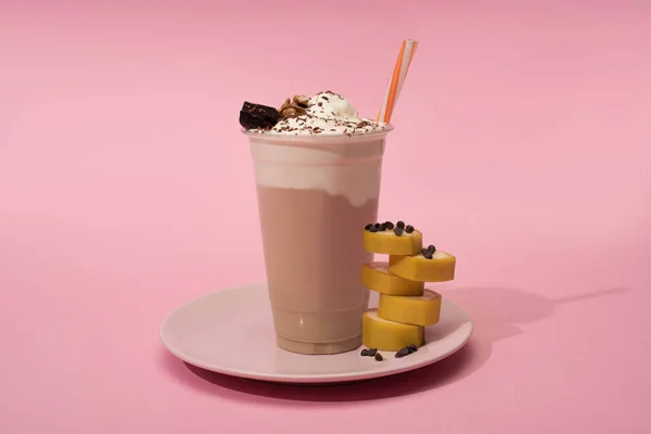 Disposable cup of milkshake with drinking straw, cut banana and chocolate chips on plate on pink background — Stock Photo