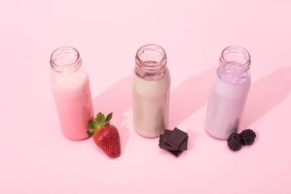 Vista de alto ângulo de garrafas de milkshakes com morango, amoras e pedaços de chocolate no fundo rosa — Fotografia de Stock