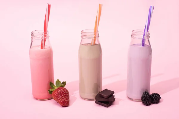 Botellas de batidos con pajitas para beber cerca de fresas, moras y trozos de chocolate sobre rosa - foto de stock