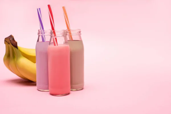 Botellas de batidos de bayas, fresas y chocolate con pajitas para beber cerca de plátanos sobre fondo rosa - foto de stock