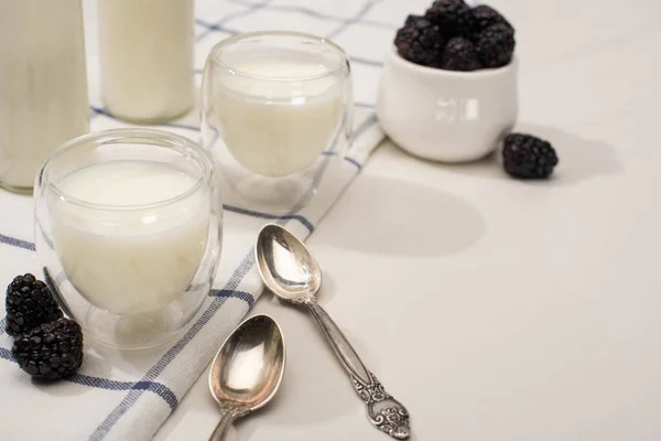 Enfoque selectivo de botellas y vasos de yogur casero con moras sobre tela cerca de cucharaditas sobre blanco - foto de stock