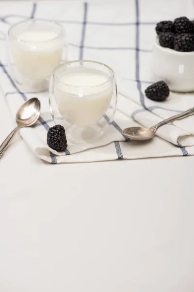 Selective focus of glasses of homemade yogurt, teaspoons and sugar bowl with blackberries on cloth on white — Stock Photo