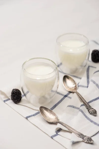 Vista en ángulo alto de vasos de yogur casero, cucharaditas y moras sobre tela sobre fondo blanco - foto de stock