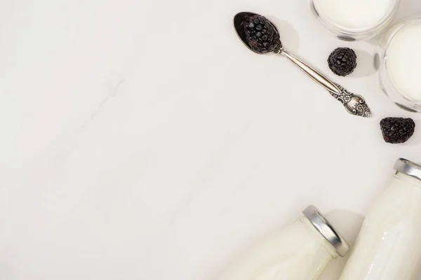 Top view of bottles and glasses of homemade yogurt and teaspoon with blackberries on white background — Stock Photo