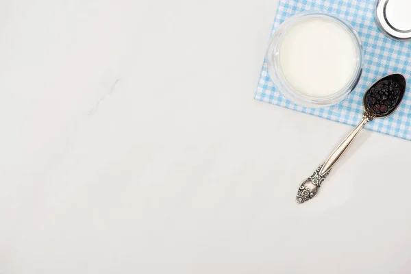 Top view of glass of homemade yogurt and teaspoon with blackberry on plaid fabric on white background — Stock Photo