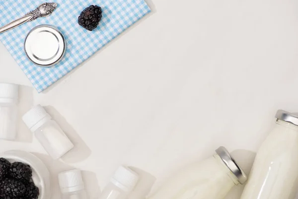 Вид сверху на ткань с крышкой и ежевикой рядом с контейнерами со стартерами и бутылками домашнего йогурта на белом — стоковое фото