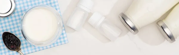 Top view of bottles and glass of yogurt, teaspoon with blackberry on cloth near containers with starter cultures on white, panoramic shot — Stock Photo