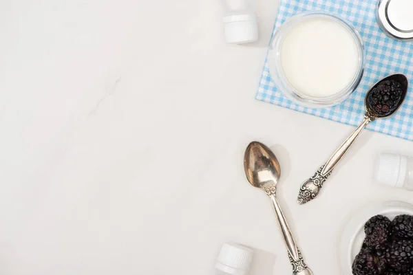 Vista dall'alto di vetro di yogurt su stoffa, cucchiaini, more e contenitori con colture di antipasto su bianco — Foto stock