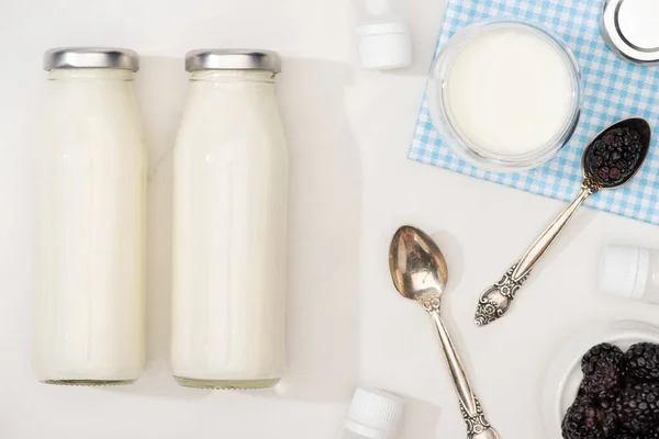 Vista dall'alto di bottiglie e bicchiere di yogurt su stoffa, cucchiaini, more e contenitori con colture di antipasto su bianco — Foto stock