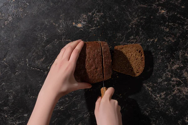 Vue recadrée de la femme coupant le pain brun avec couteau sur la surface noire de pierre — Photo de stock