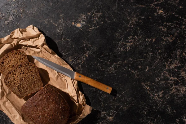 Top view of fresh cut loaf of brown bread with knife on paper on stone black surface — Stock Photo