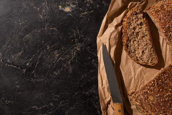 Vue du dessus du pain coupé à grains entiers sur papier près d'un couteau sur une surface noire en pierre — Photo de stock