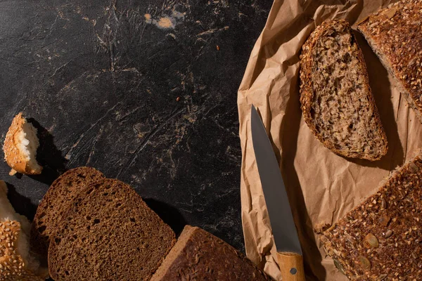 Vista superior del pan de grano entero cortado en papel cerca del cuchillo en la superficie de piedra negro - foto de stock