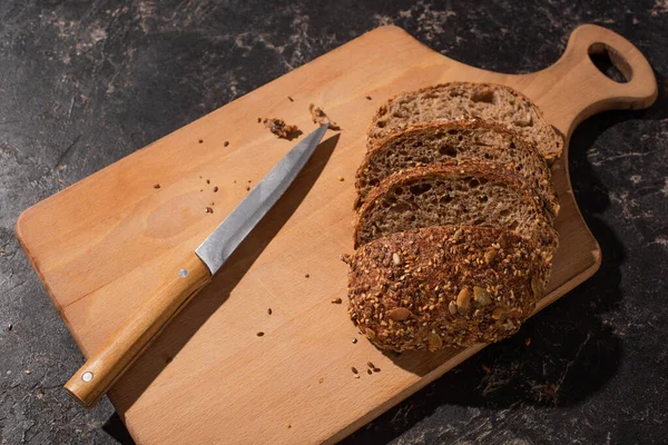 Cortar el pan de grano entero en la tabla de cortar cerca del cuchillo en la superficie de piedra negro - foto de stock