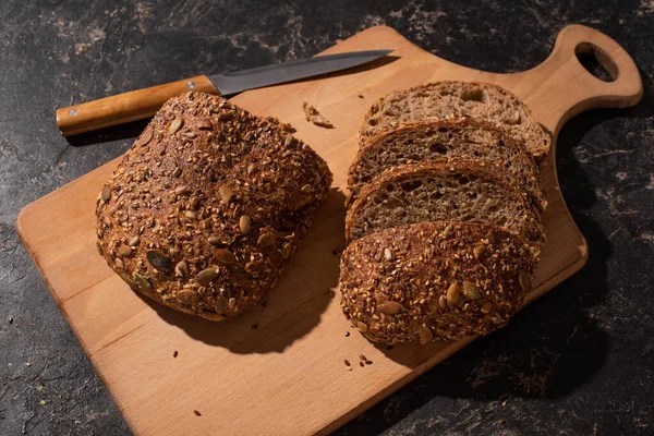 Cortar pão de grão inteiro na placa de corte perto da faca na superfície preta de pedra — Fotografia de Stock